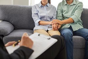Cropped shot of psychologist taking notes on clipboard during family therapy session with young married couple. Husband and wife sitting on couch, holding hands, trying to solve relationship problem.