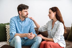 Support, trust and understanding in marriage. Young husband sitting on sofa at home and talking about problems, wife listening carefully and hugging man. Overcome family difficulties together.